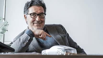 Porträtaufnahme einer Person im Sakko an einem Schreibtisch mit einem Automodell im Vordergrund.